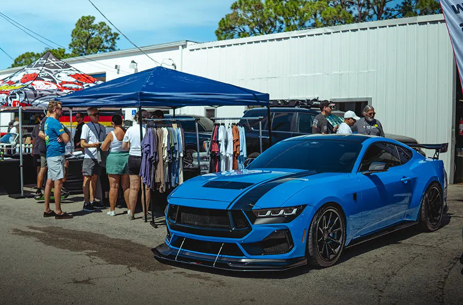 A blue Ford Mustang Dark Horse parked outside the Superior Autohaus garage in Bonita Springs, Florida, attending one of their local events. A vendor is set up nearby selling t-shirts. 