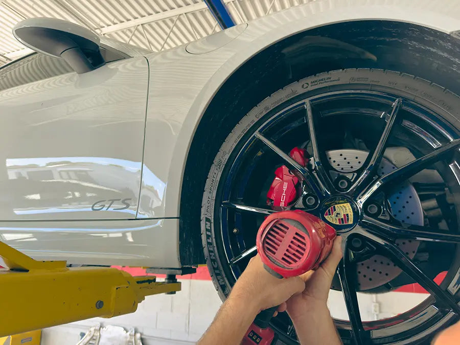 A Superior Autohaus mechanic using an impact wrench on the lugnuts of a white Porsche 911 GTS up on a lift. 